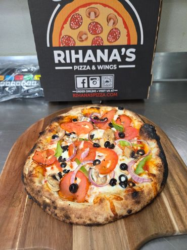 A freshly baked veggie pizza on a wooden board with a Rihana's pizza box in the background.