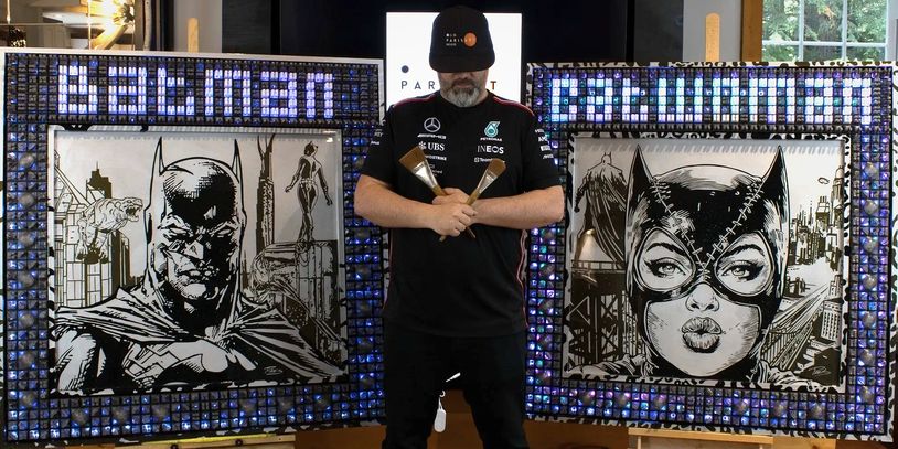 Artist posing with Batman and Catwoman paintings, holding brushes.