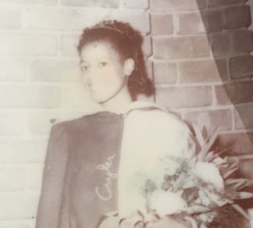 Vintage photo of a young woman with a sash and bouquet in front of a brick wall.