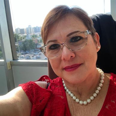 A woman in red dress and pearl necklace smiling indoors by a window.