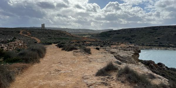A rocky coastal path under a cloudy sky with distant hills and water.