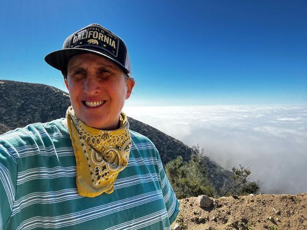 Person smiling with a California cap on a sunny mountain hike above the clouds.