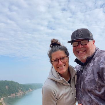 Smiling couple posing outdoors with a scenic river and hills in the background.