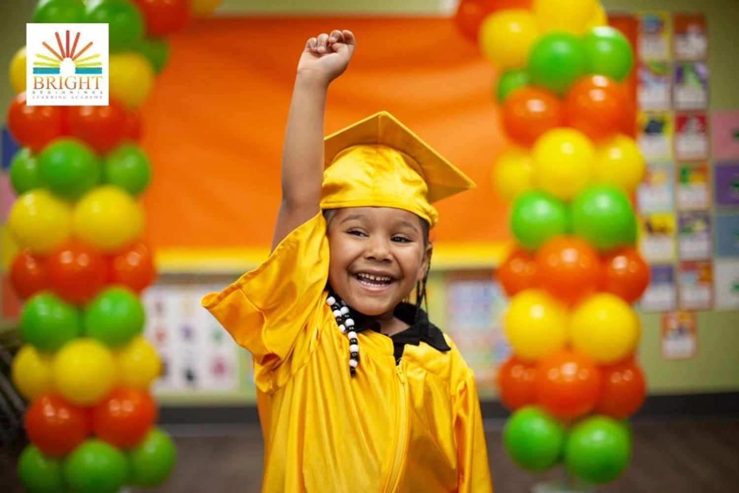 Smiling child in yellow graduation gown raising fist joyfully amid colorful balloon decorations.