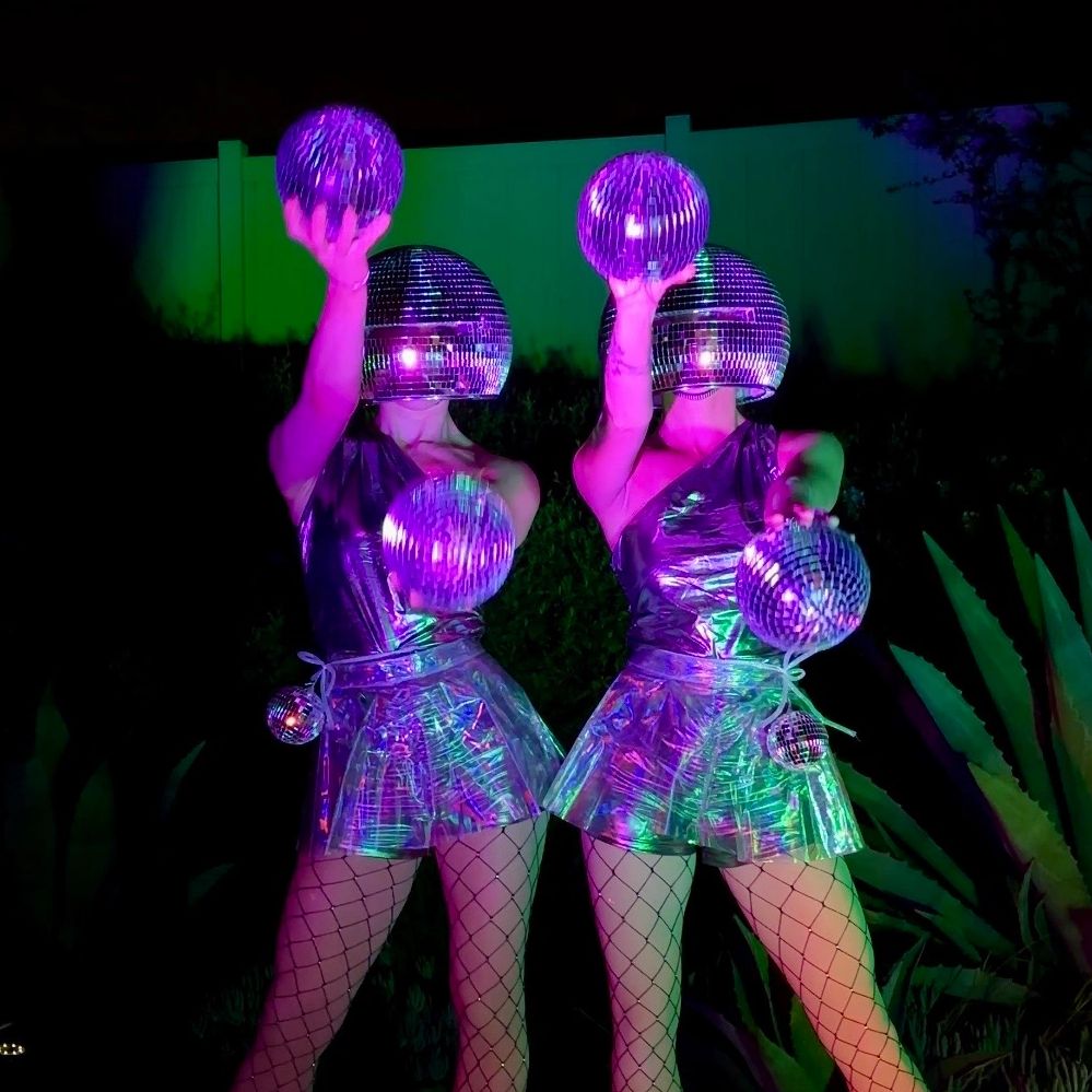 Two people in disco ball helmets and shiny outfits holding disco balls.