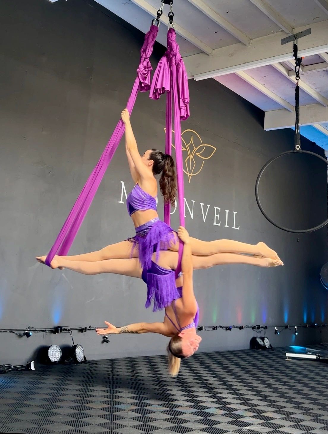 Two acrobats in purple perform aerial silk at a studio.