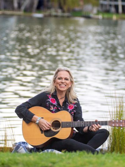 Woman sitting by the water, playing an acoustic guitar and smiling.