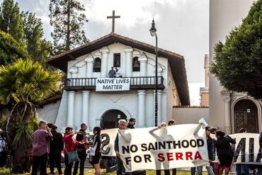 Protesters hold banners in front of a church advocating Native lives and opposing sainthood for Serra.