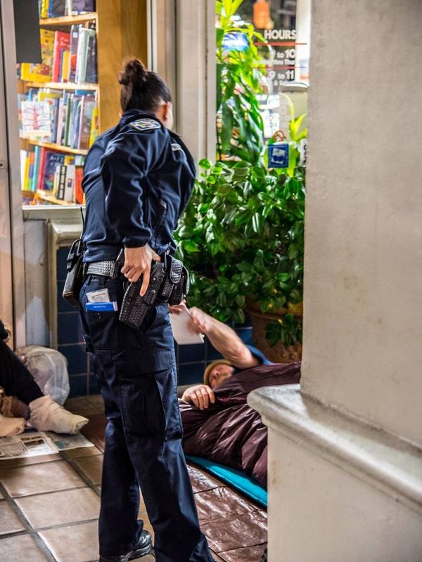 Police officer holding their gun while issuing a citation to a homeless person lying on the ground outside a building.