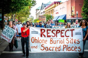 Protesters march holding signs advocating respect for Ohlone burial sites and sacred places.