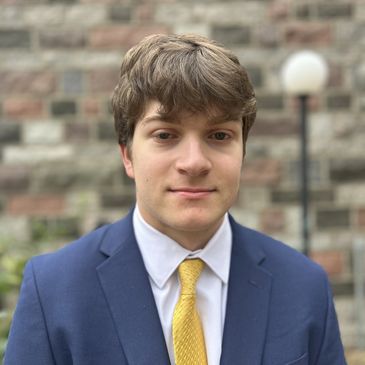 Young man in a blue suit and yellow tie with a brick wall background.