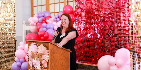 Woman speaking at a colorful balloon-decorated event podium.