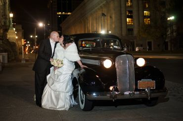 Bride and groom kiss beside a vintage car at night in a city.