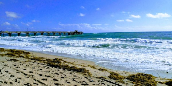 Sunny beach with waves and a pier extending into the ocean under a blue sky.