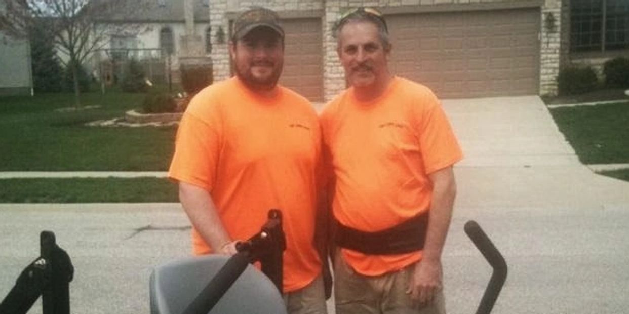 Two men in orange shirts stand behind a red Exmark Lazer Z lawn mower.