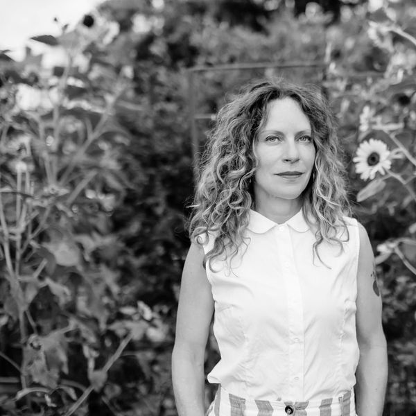 Woman with curly hair stands in a garden surrounded by sunflowers.