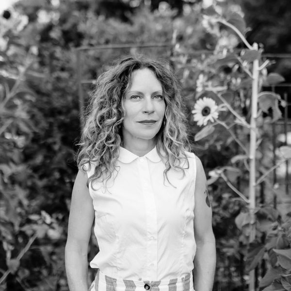 Woman with curly hair stands in a garden surrounded by sunflowers.
