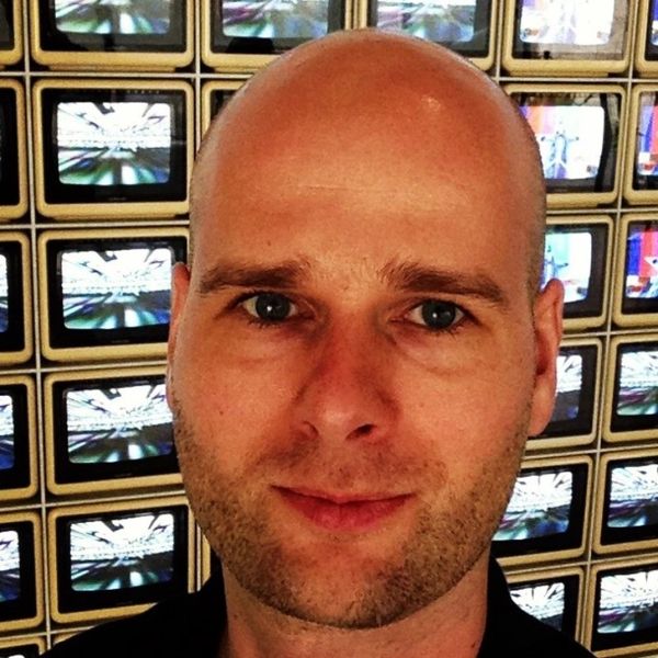 Close-up of a bald man with a slight smile in front of multiple small TV screens.