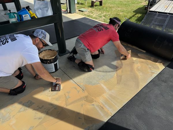 Two workers are applying adhesive to a concrete surface before laying down rubber flooring rolls.