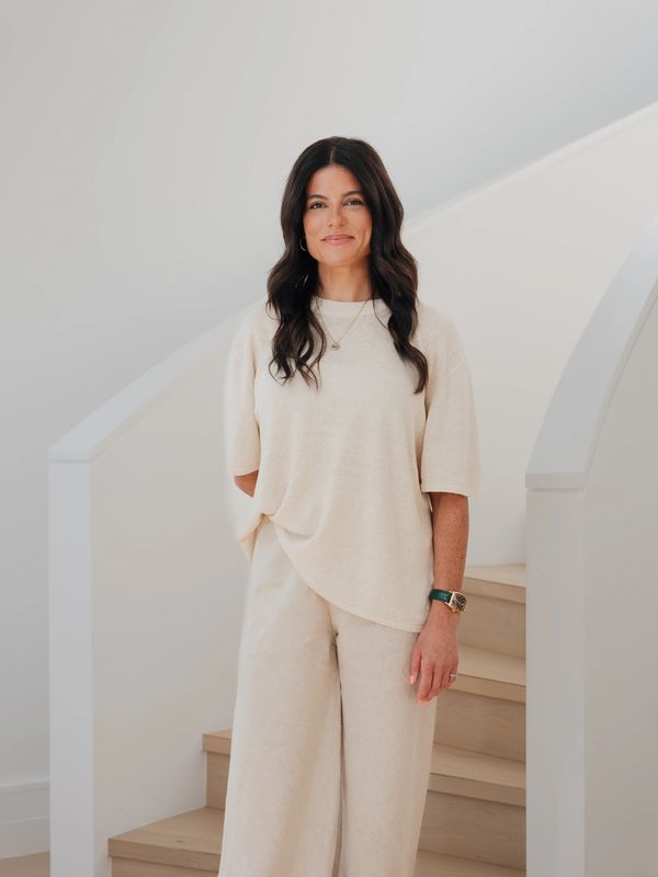 Woman with long dark hair in beige outfit standing by wooden stairs.