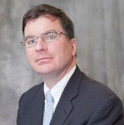 A dark-haired distinguished gentleman in a grey suit and glasses stares into the camera.