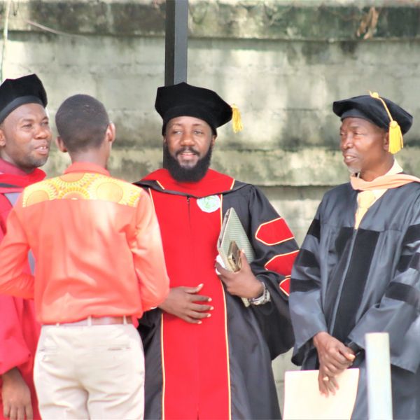 Graduates in academic regalia converse with a casually dressed man outdoors.