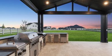 Modern outdoor kitchen with stainless steel appliances under a covered patio at sunset.