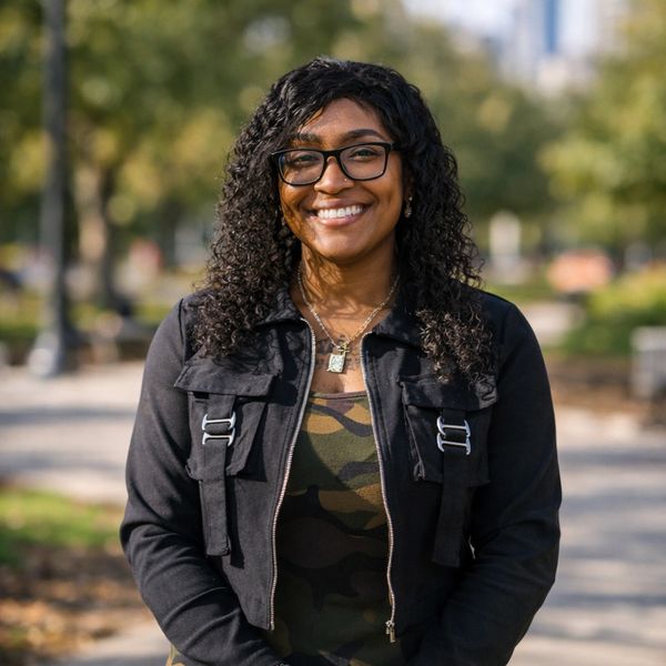 Smiling woman with curly hair wearing glasses and a black jacket outdoors.