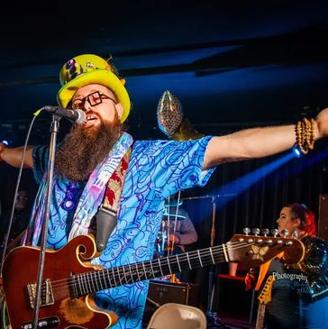 Bearded musician with a colorful hat and guitar performs energetically on stage.