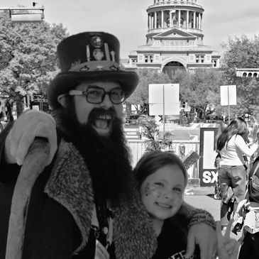 Man in top hat with beard and a smiling girl at outdoor event.