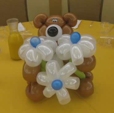Balloon sculpture of a bear holding white flowers with blue centers on a yellow table.