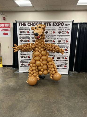 Person dressed in a detailed bear costume made of brown balloons at a chocolate expo.