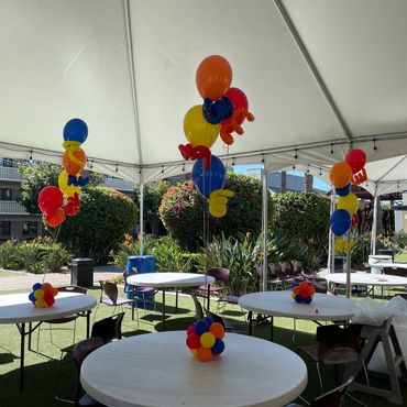 Colorful balloon centerpieces decorate tables under a white party tent outdoors.