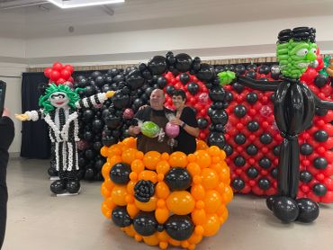 People posing with Halloween-themed balloon sculptures including a pumpkin and Frankenstein.