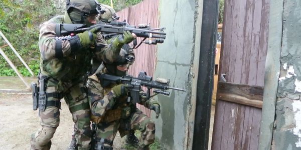 Two soldiers in camouflage aiming rifles near a wooden fence in a tactical stance.