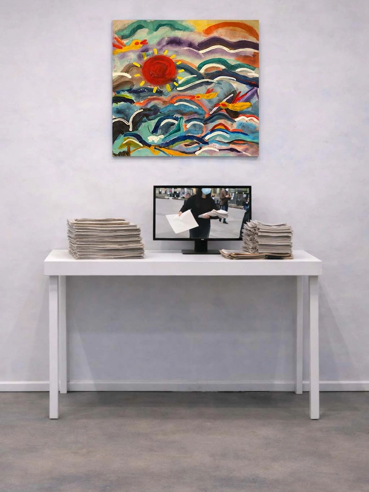 White table with stacks of newspapers and a monitor showing a person distributing papers.