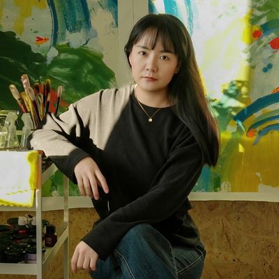 A young woman posing thoughtfully in an art studio with paintings and brushes.