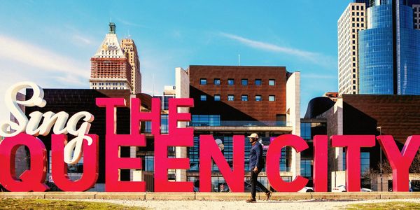 Large colorful sign reading 'Sing The Queen City' with a person walking nearby in an urban setting.