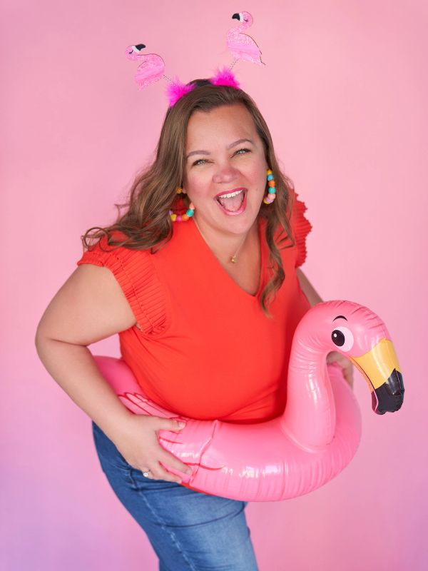 Woman joyfully posing with pink flamingo float and headband against pink background.