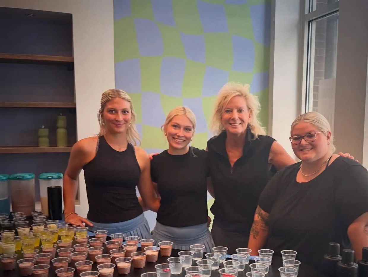 Four women posing behind a table with many small cups of drinks.