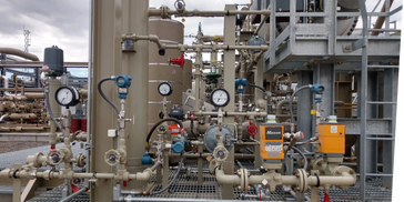 Gas processing equipment with pressure gauges, valves, and piping on metal grating platform.