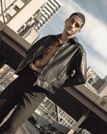 Stylish man in a black leather jacket posing outdoors in an urban setting.