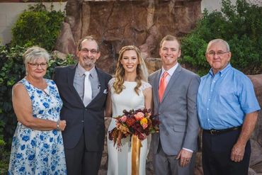 Groom wearing light grey suit with orange tie standing as newlyweds. Their fathers are also in suits