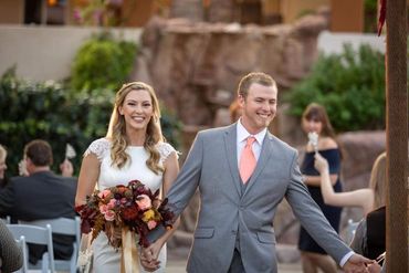 Groom wearing light grey suit with orange tie, walking his bride down the aisle at their wedding