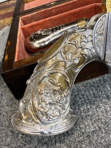 Intricately engraved silver pistol grip with a wooden box and powder flask in the background.