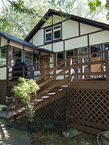 A rustic house with a brown wooden deck and stairs surrounded by greenery.