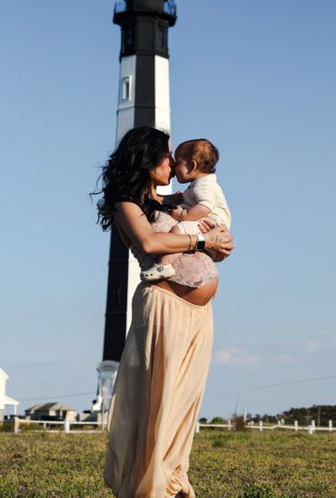 Pregnant woman lovingly holding and kissing her baby outdoors by a lighthouse.