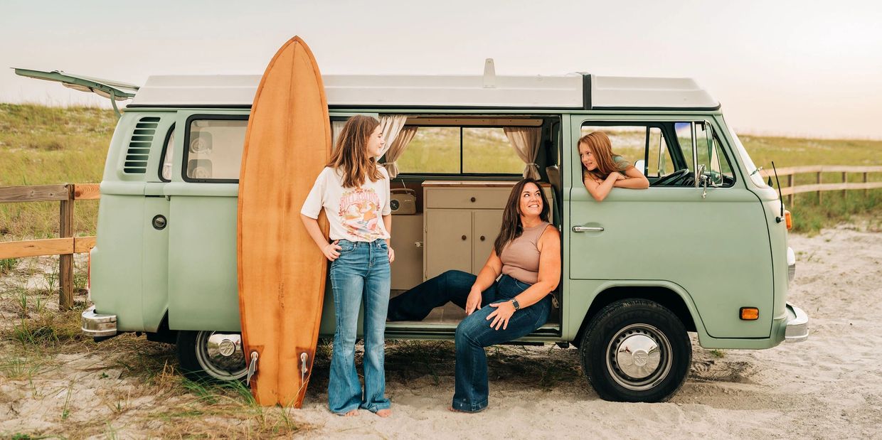 Three women with a surfboard near a vintage van on the beach.
