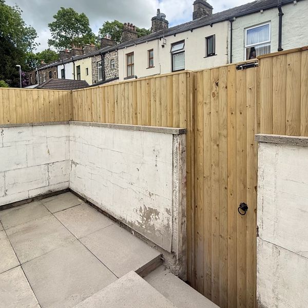 New wooden fence and gate enclosing a small backyard area beside older stone houses.