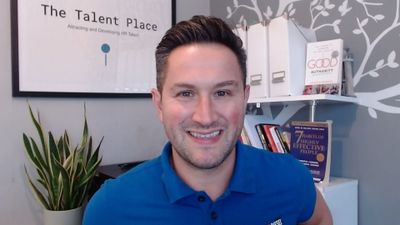 Smiling man in blue polo shirt sitting in a home office with books and plant.
